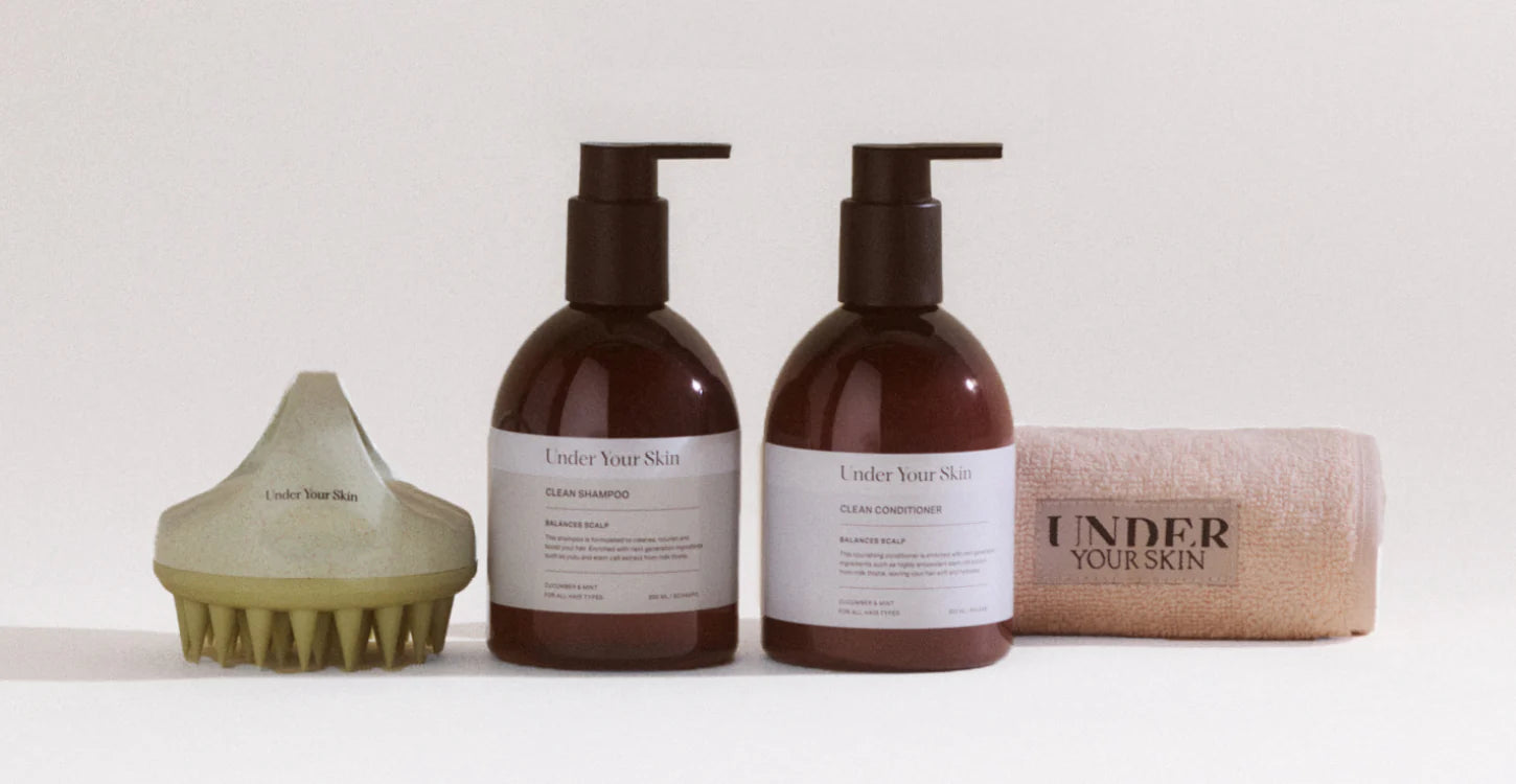 Under Your Skin scalp brush, schampo-, balsamflaskor och märkt handduk på vit bakgrund