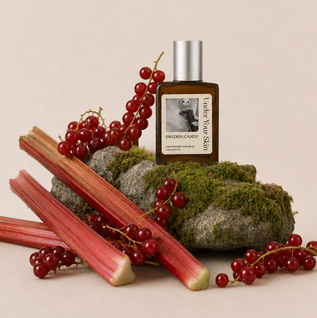 A small amber glass bottle labeled 'Swedish Candy" with a silver cap, styled on mossy rocks with red rhubarb stalks and red berries against a warm beige background.